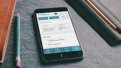 Photo of a smartphone lying on a table along with pencils and a notebook. The screen shows Taskly's dashboard.